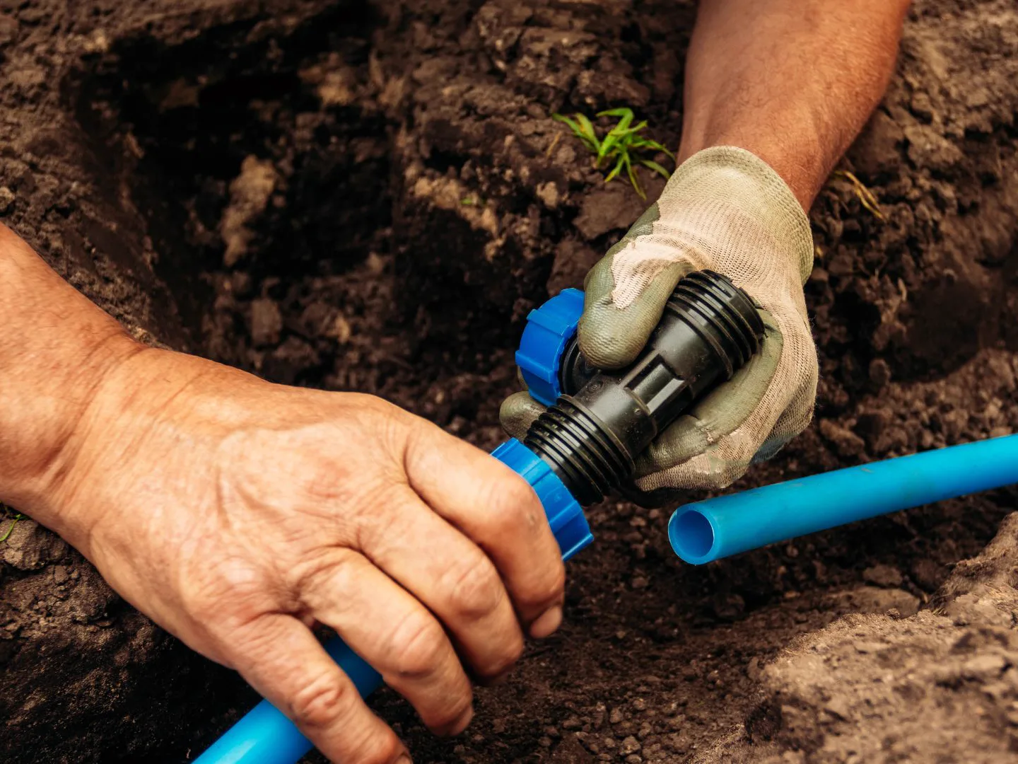 Engineer's gloved hand joining a black compression fitting to blue water pipes in a soil trench